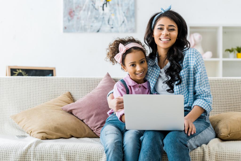 sonriente afroamericano mamá y su hija usando el ordenador portátil mientras está sentado en el sofá
 - Foto, imagen