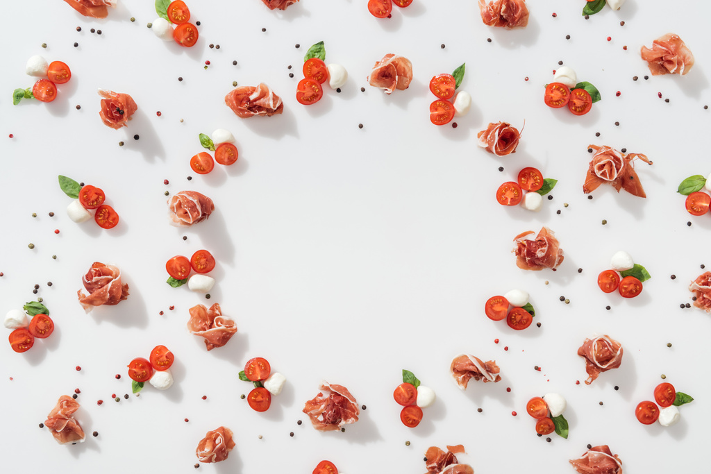 flat lay of prosciutto near red cherry tomatoes, mozzarella cheese, green basil leaves and peppercorns on white background - Photo, Image
