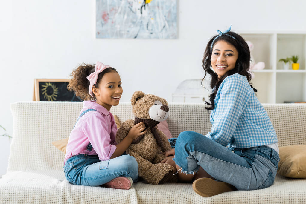 cute african american child holding teddy bear while sitting on sofa with mother - Photo, Image