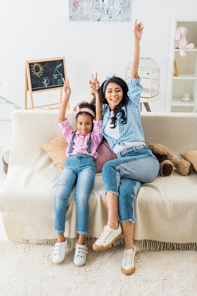 cheerful african american mom and daughter showing victory signs while sitting on sofa at home - Photo, Image