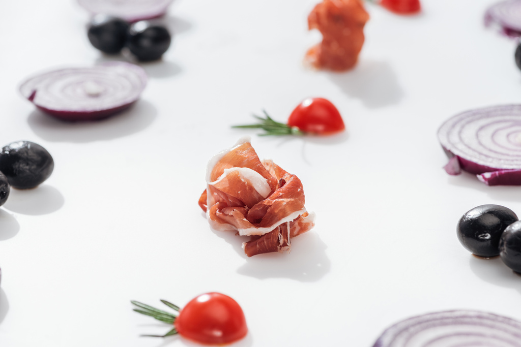 selective focus of tasty prosciutto near cherry tomatoes with rosemary twigs near red onion rings and black olives on white background - Photo, Image