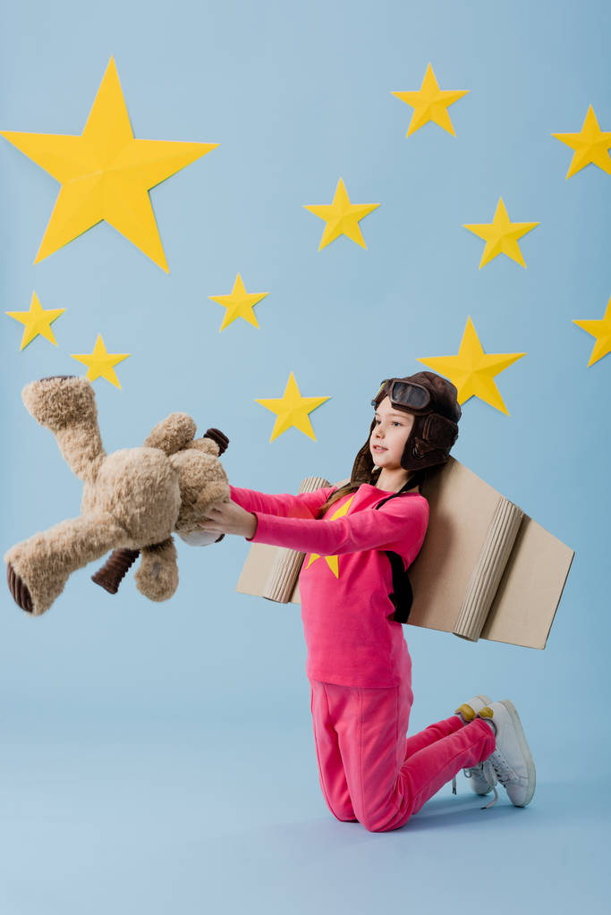 Kid met kartonnen vleugels staande op de knieën en houden van teddybeer op blauwe achtergrond van de sterrenhemel - Foto, afbeelding