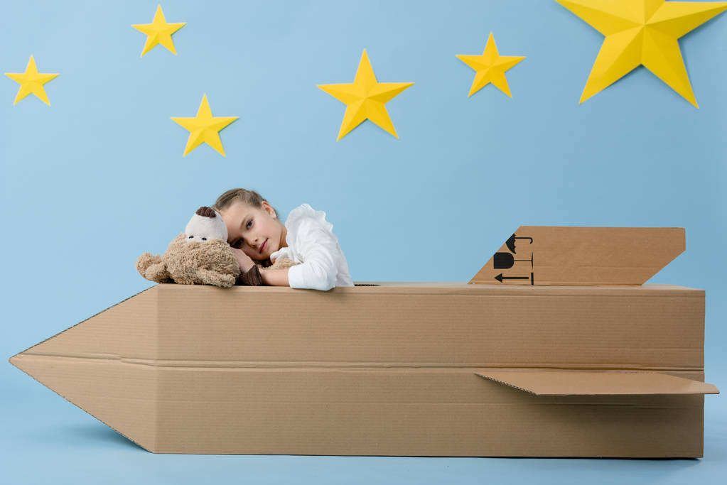 Preteen kid with teddy bear sitting in cardboard rocket on blue starry background - Photo, Image
