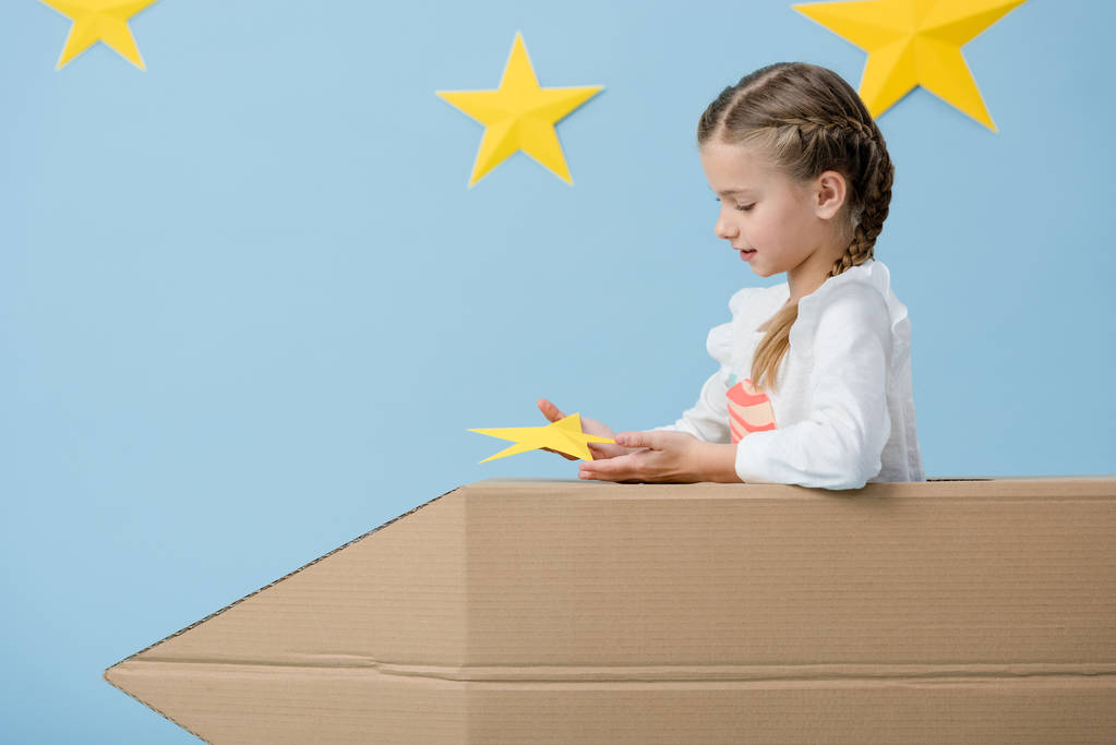 Criança com tranças sentadas em foguete de papelão e segurando estrela amarela no fundo estrelado azul
 - Foto, Imagem