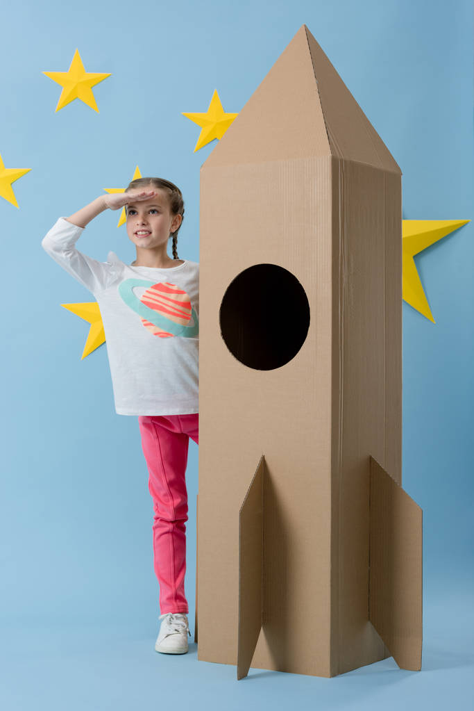 fröhliches Kind, das neben einer Papprakete steht und auf blauem Sternenhintergrund wegschaut - Foto, Bild