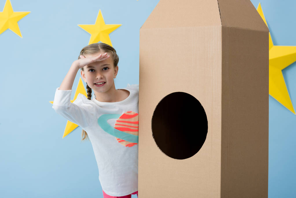Χαμογελαστό παιδί στέκεται κοντά πύραυλος από χαρτόνι και αναζητούν μακριά σε μπλε φόντο έναστρο - Φωτογραφία, εικόνα