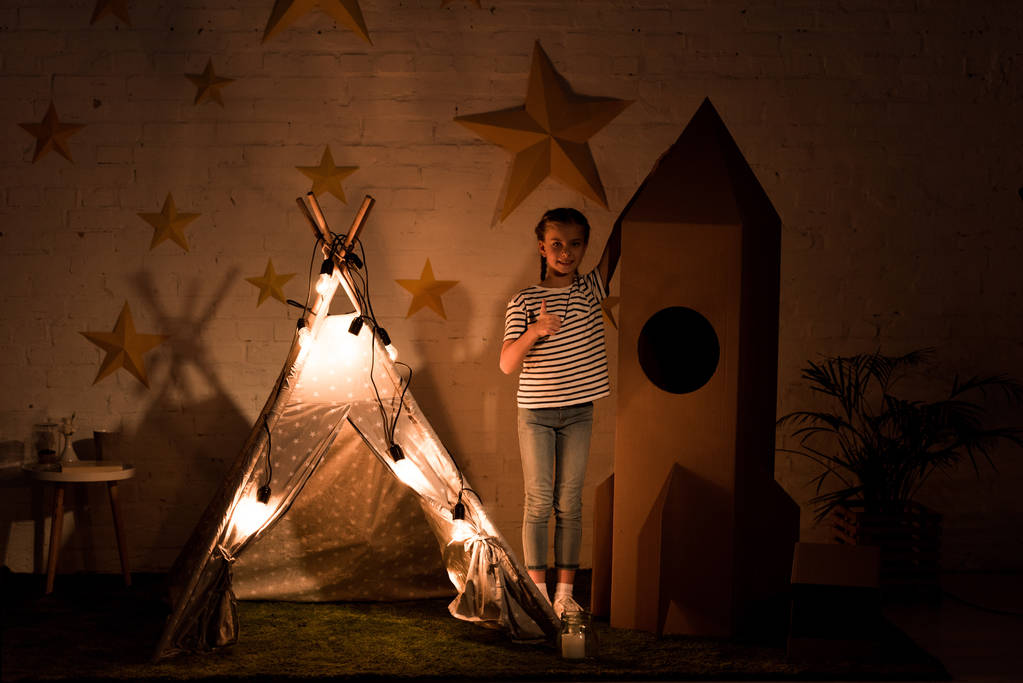 Criança de pé entre wigwam e foguete de papelão no quarto escuro
 - Foto, Imagem