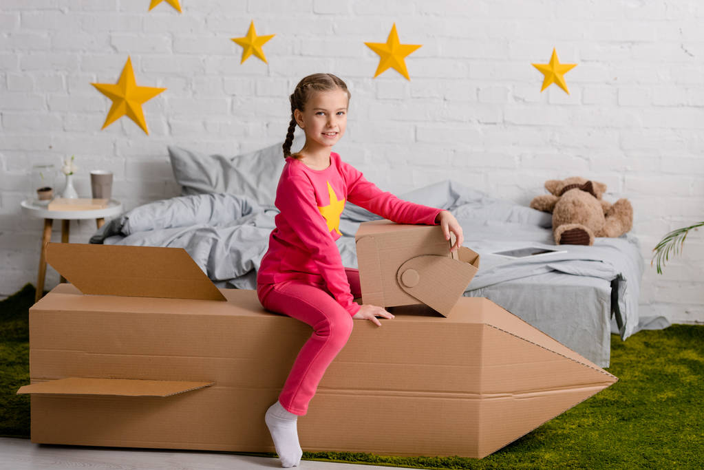 Vreugdevolle kind met helm zittend op kartonnen raket in slaapkamer - Foto, afbeelding