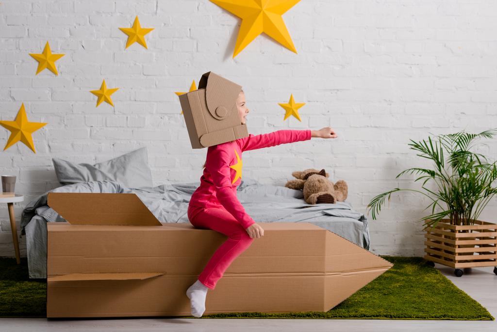 Ragazzo divertente in casco di cartone seduto su un razzo con la mano in camera da letto
 - Foto, immagini