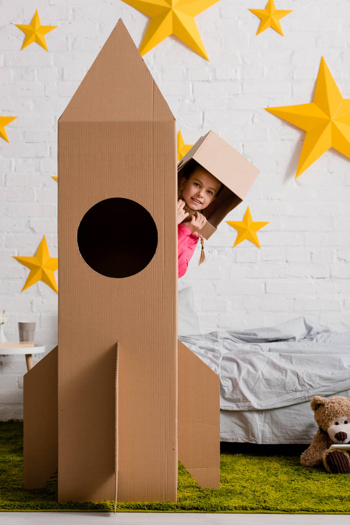 Unbekümmertes Kind mit Papphelm steht hinter Rakete im Schlafzimmer - Foto, Bild