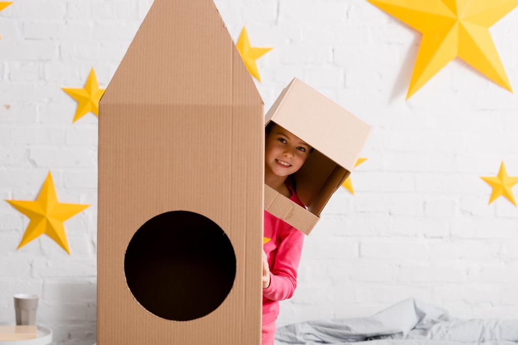 Смешной ребенок в картонном шлеме стоит рядом с ракетой в спальне
 - Фото, изображение