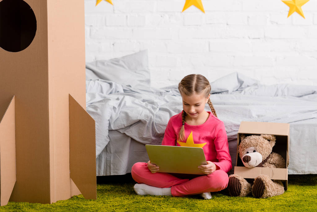 Blij kind zittend op een tapijt met teddybeer en met behulp van laptop - Foto, afbeelding
