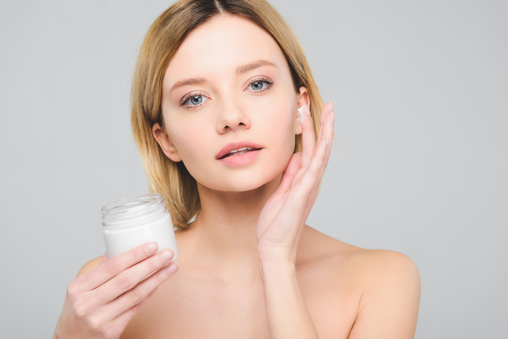 attractive girl applying cosmetic cream on face and holding plastic container isolated on grey - Photo, Image