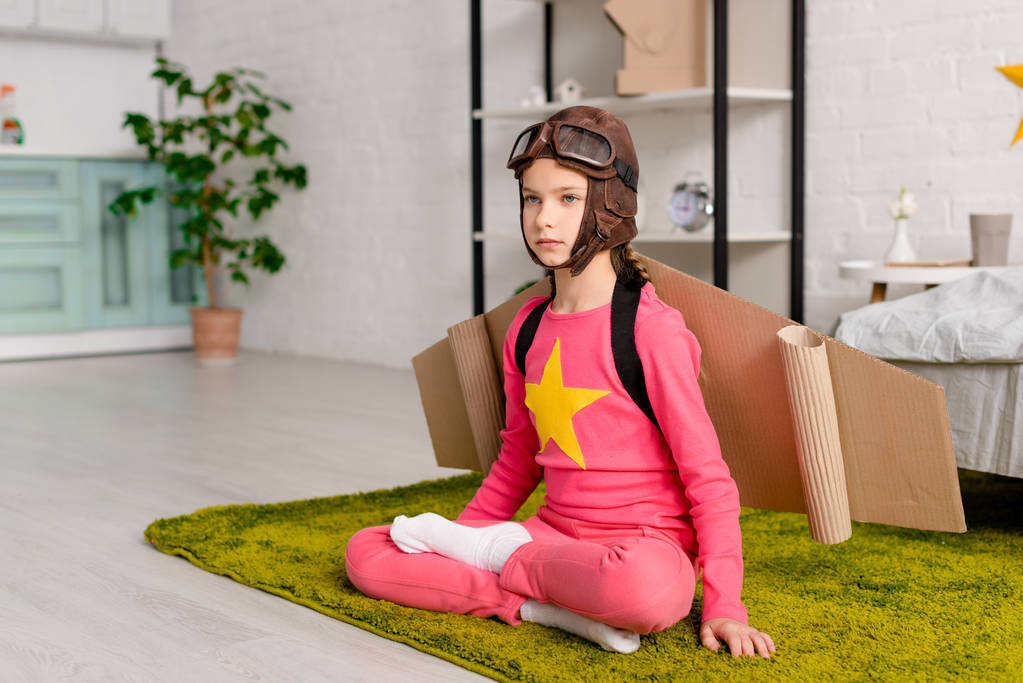 Серйозна дитина з картонними крилами, що сидить у позі лотоса на килимі
 - Фото, зображення