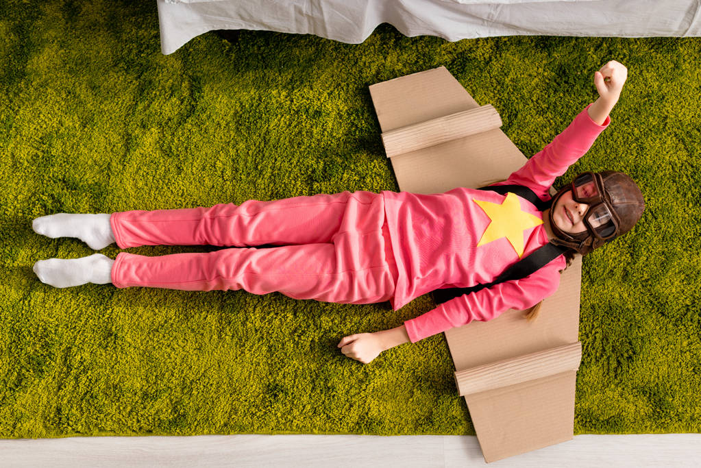 Vista superior de niño divertido con alas de cartón acostado en la alfombra con el puño hacia arriba
 - Foto, Imagen