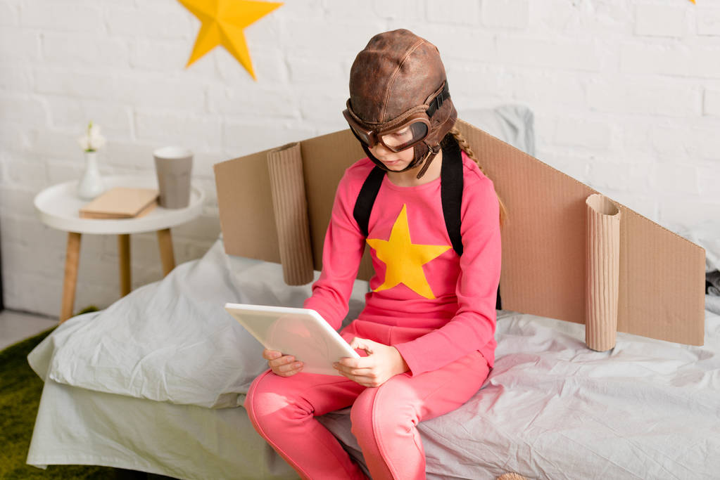 Παιδί με ροζ ρούχα και κράνος πτήση χρησιμοποιώντας ψηφιακό tablet ενώ κάθεται στο κρεβάτι - Φωτογραφία, εικόνα