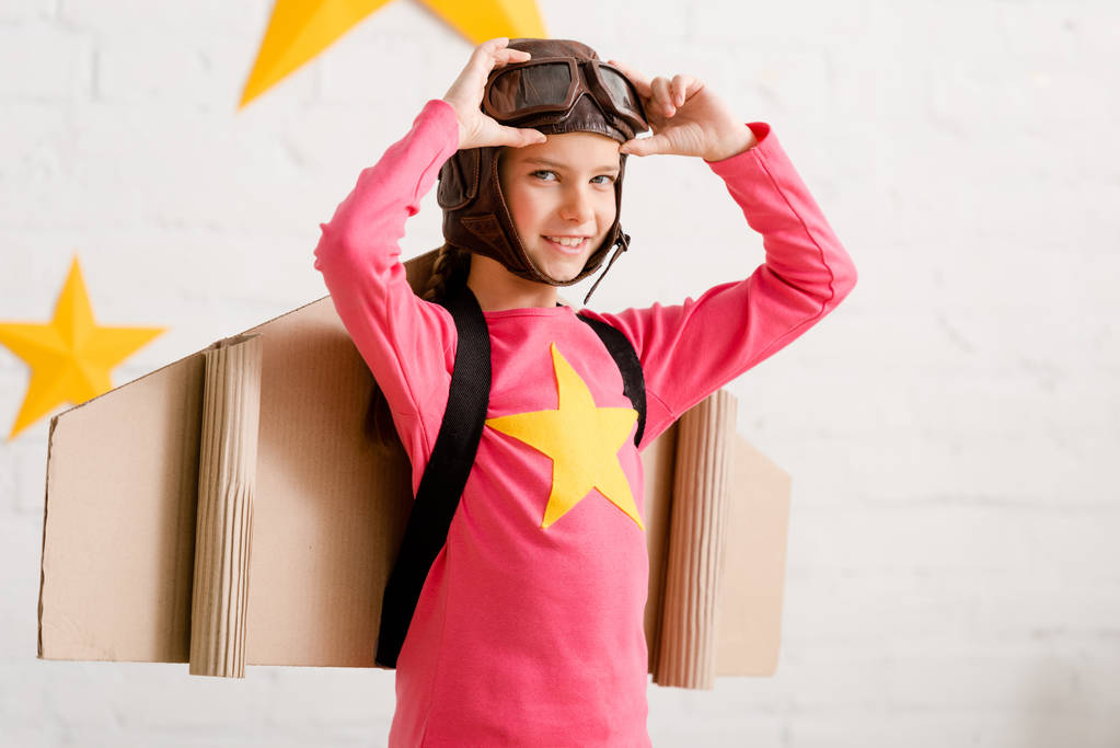 Pleased child with cardboard wings touching goggles with smile - Photo, Image
