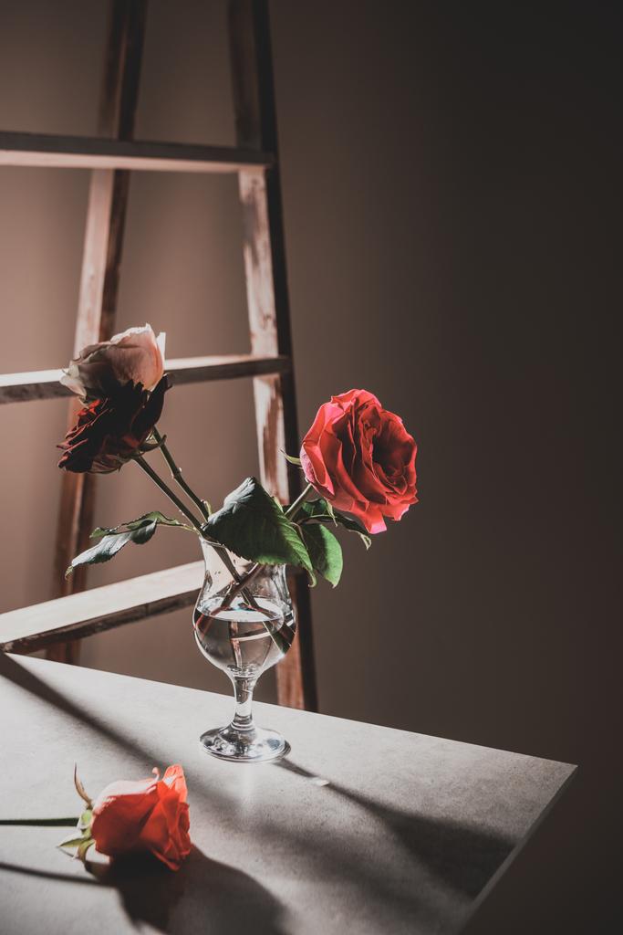 rote Rosenblüten im Weinglas auf Steintisch mit Holzleiter auf Hintergrund - Foto, Bild