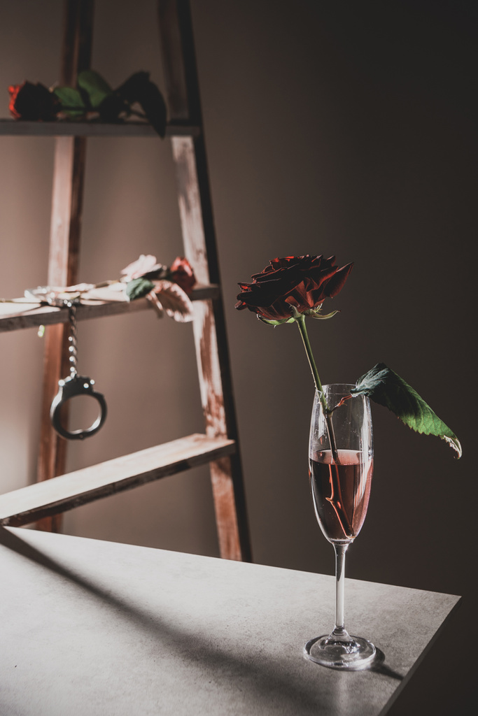 цветы красной розы в бокале шампанского на каменном столе с деревянной лестницей и металлическими наручниками на заднем плане
 - Фото, изображение