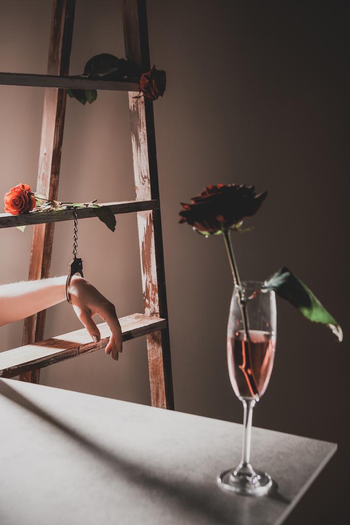 flores de rosa roja en copa de champán en mesa de piedra con escalera de madera y mujer en esposas de metal en el fondo
 - Foto, imagen