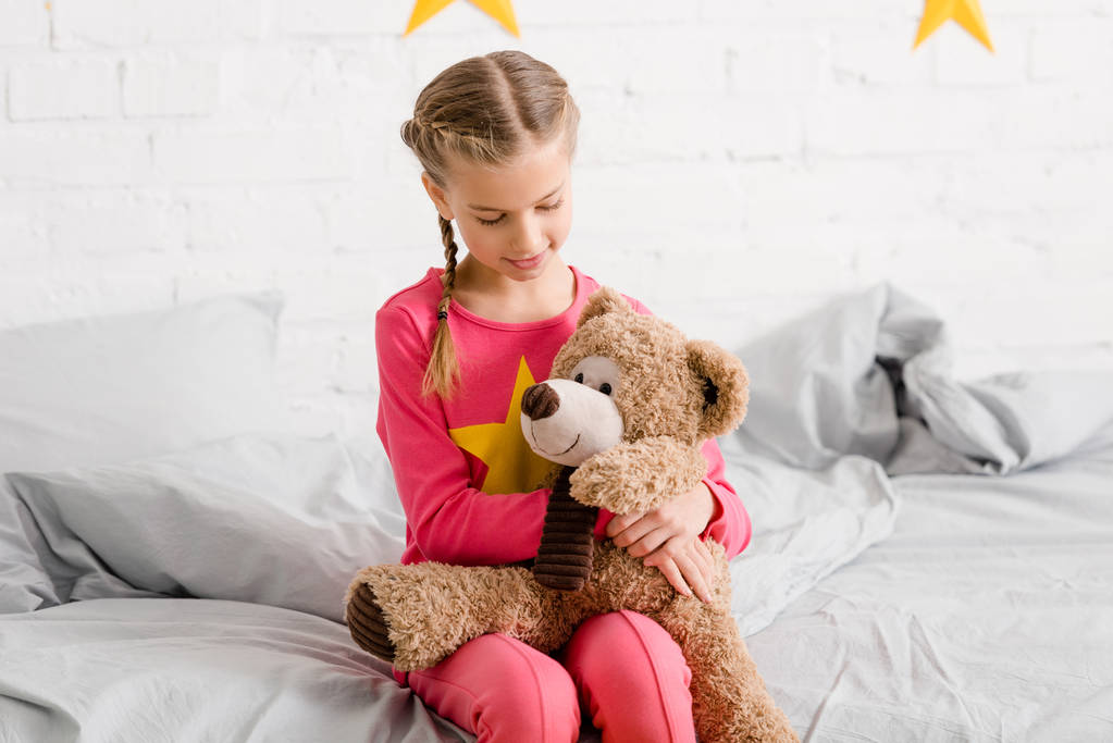 entzückendes Kind sitzt auf dem Bett und hält Teddybär auf Knien - Foto, Bild