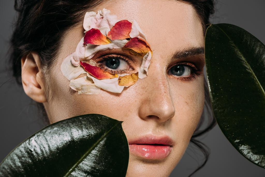 όμορφο κορίτσι με floral πέταλα γύρω από τα μάτια κρατώντας φύλλα που απομονώνονται σε γκρι - Φωτογραφία, εικόνα