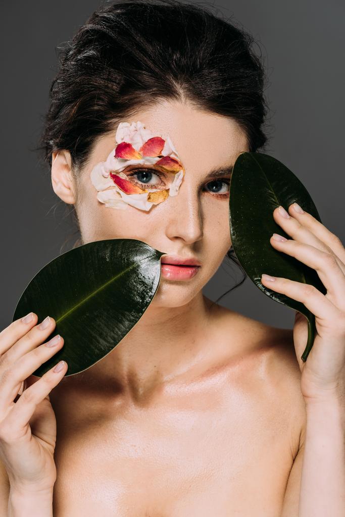 piękna młoda kobieta z płatkami kwiatów wokół oczu, trzymając liści na szarym tle - Zdjęcie, obraz