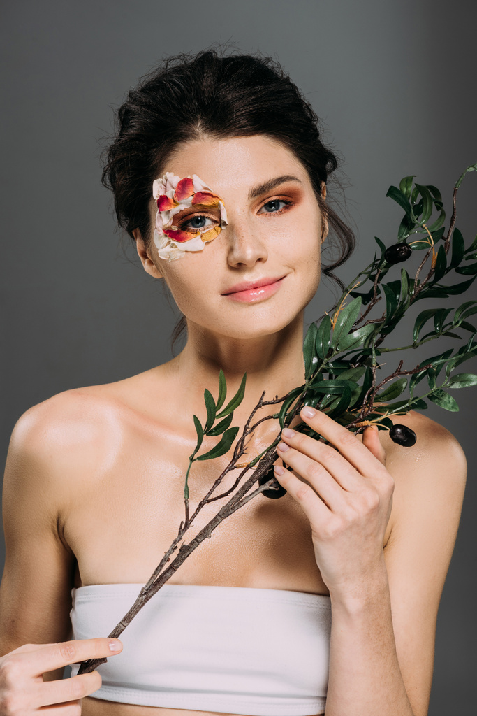 tender smiling girl with petals on face holding green branch isolated on grey - Photo, Image