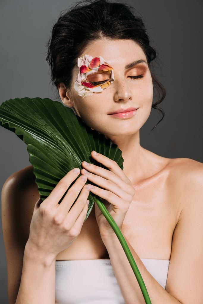 мрійлива жінка з закритими очима і квітковими пелюстками на обличчі тримає зелений лист ізольовано на сірому
 - Фото, зображення