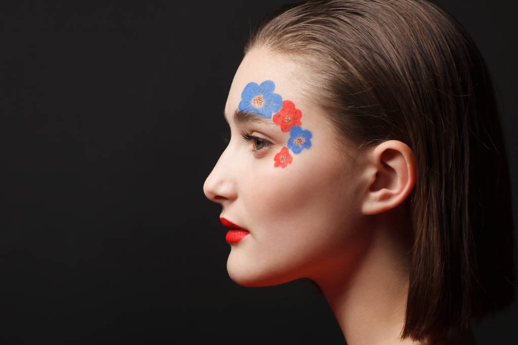 Η φωτογραφία ενός νεαρού κοριτσιού που με το makeup σε μορφή λουλούδια σε κόκκινο και μπλε. Μαύρο φόντο - Φωτογραφία, εικόνα