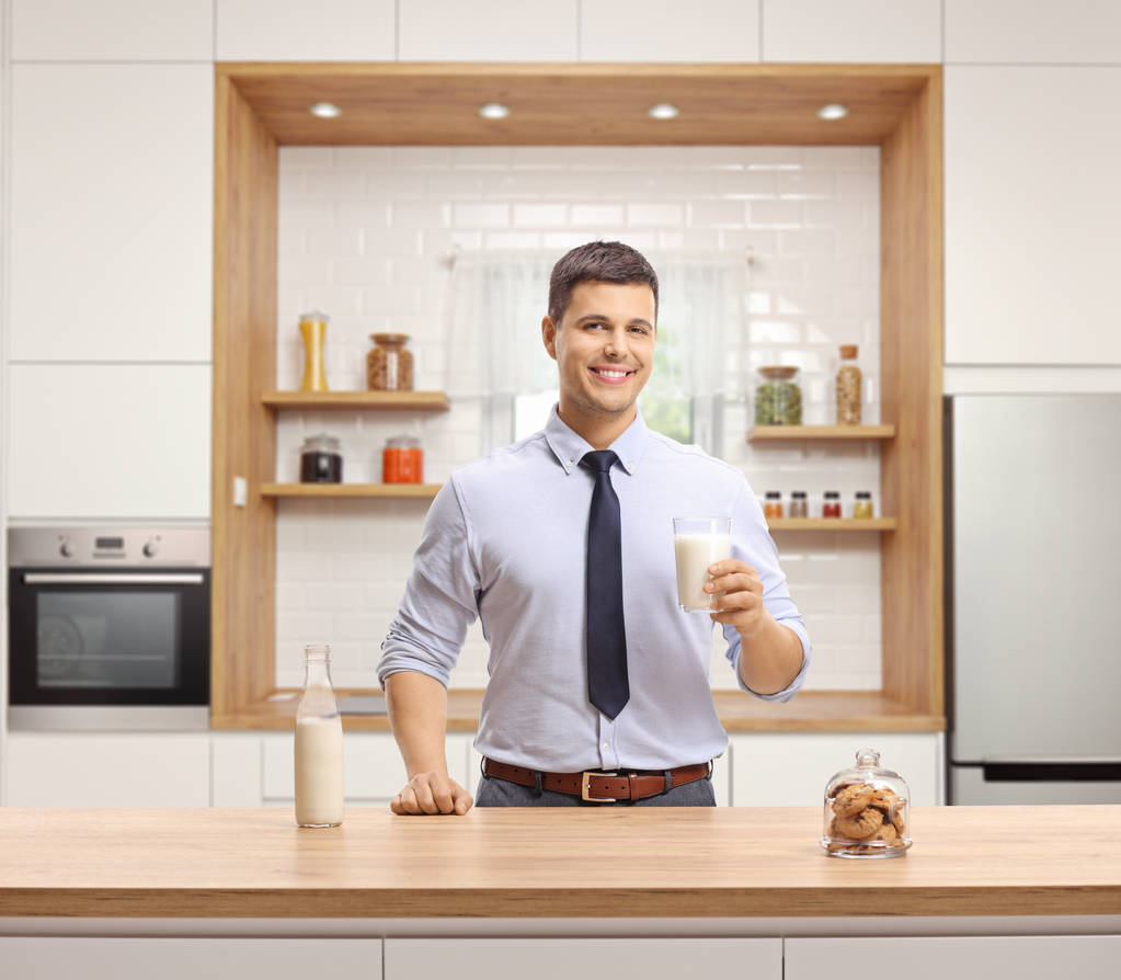 Młody człowiek w ubrania formalne trzymając szklankę mleka i stojąc w nowoczesnej kuchni - Zdjęcie, obraz