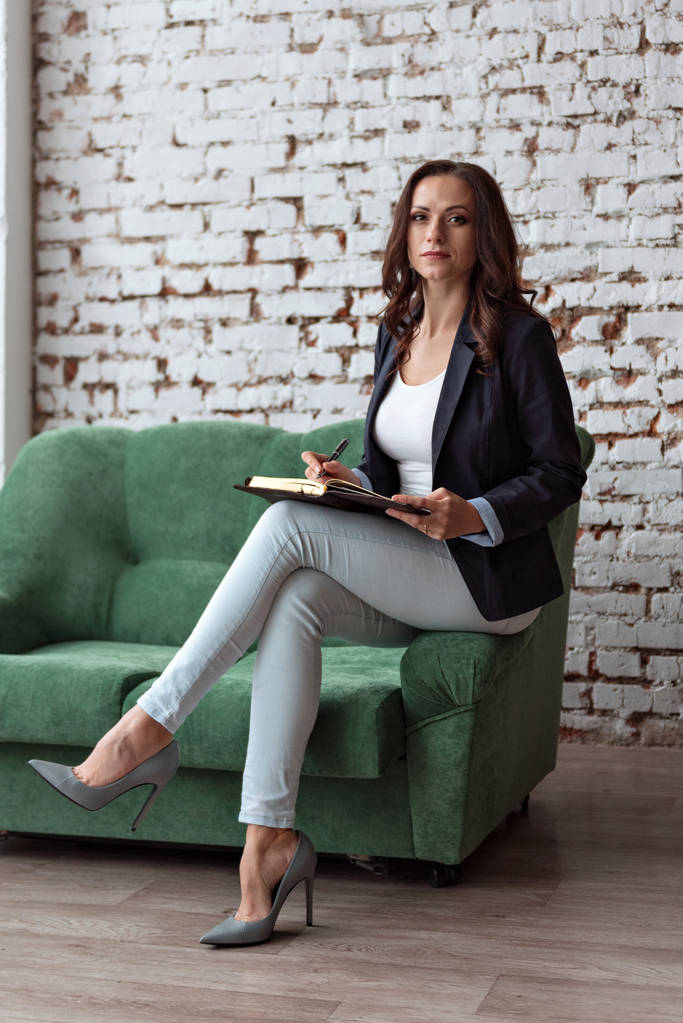 retrato de mujer entrenador de negocios sentado en el estante del sofá sosteniendo un cuaderno mirando directamente en traje de estilo de vida
 - Foto, Imagen