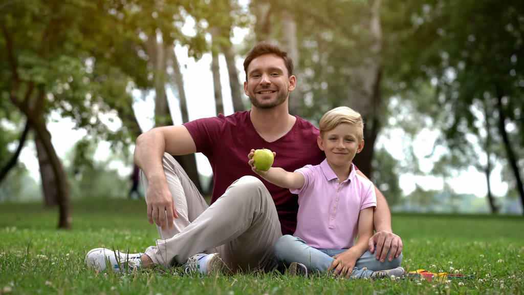 カメラ、健康食品に新鮮なリンゴを見せている公園で父親と座っている少年 - 写真・画像