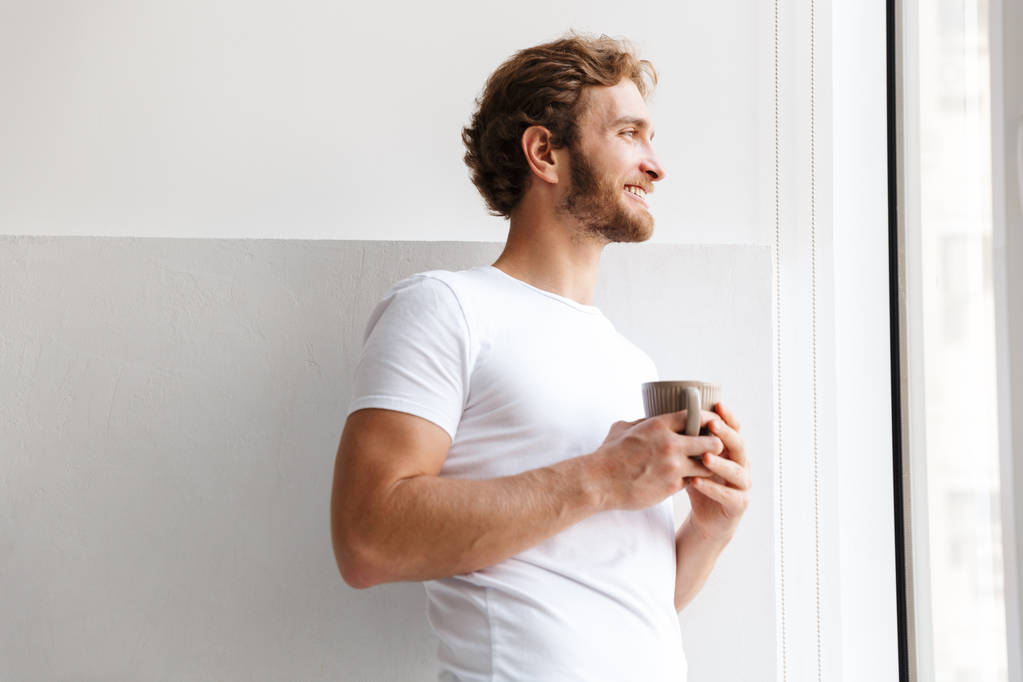 Jeune homme souriant tenant une tasse de thé, debout à la fenêtre à la maison
 - Photo, image
