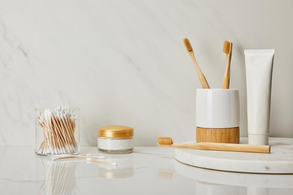 зубная паста в тюбике, держатель, бамбуковые зубные щетки, косметический крем и ушные палочки в стекле на белом мраморном фоне
 - Фото, изображение