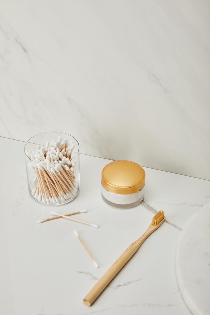 bâtons d'oreille en verre, crème cosmétique et brosse à dents en bambou sur fond de marbre blanc
 - Photo, image