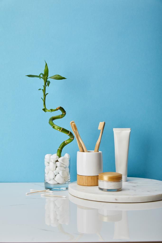 cepillos de dientes en soporte, crema cosmética y pasta de dientes en tubo sobre tabla redonda cerca del tallo de bambú verde en jarrón sobre fondo azul
 - Foto, Imagen