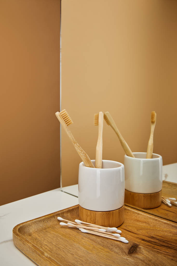 Brett mit Bambuszahnbürsten im Halter und Ohrstöpseln neben Spiegel auf weißem Tisch und beigem Hintergrund - Foto, Bild