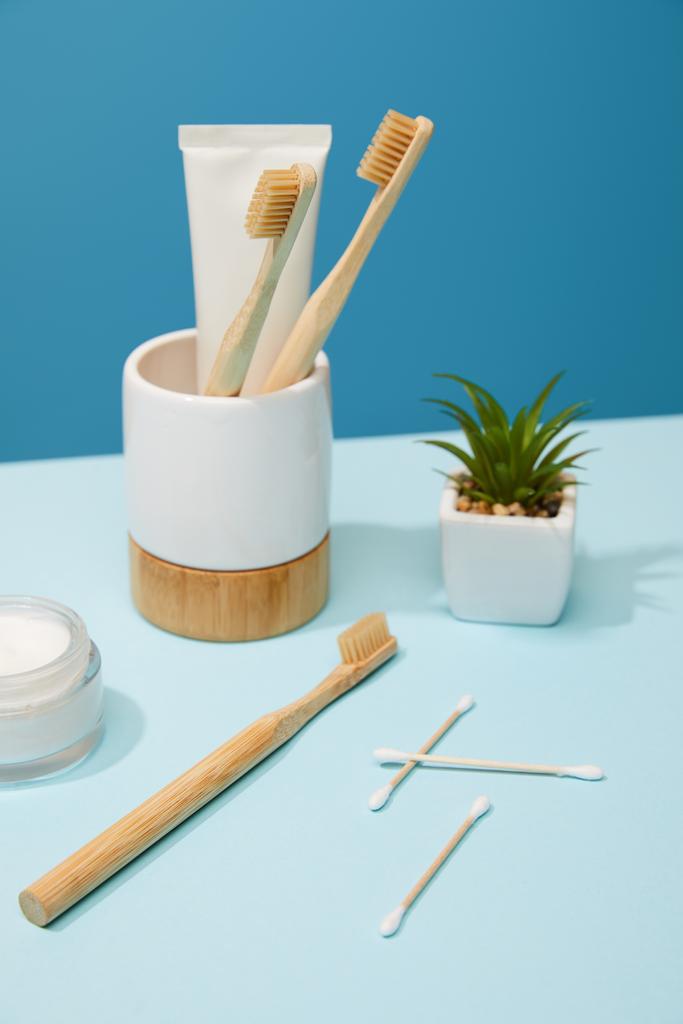 držák s zubní pastu v trubici a bambusové kartáčky, kosmetický krém a závod v hrnci na stůl a modré pozadí - Fotografie, Obrázek