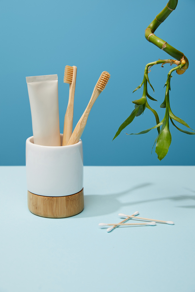 holder with bamboo toothbrushes, toothpaste in tube, ear sticks and bamboo stem on table and blue background - Photo, Image