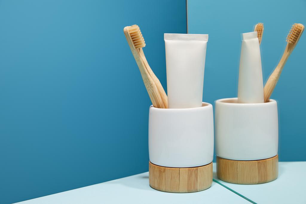 holder with bamboo toothbrushes, toothpaste in tube near mirror on table and blue background - Photo, Image