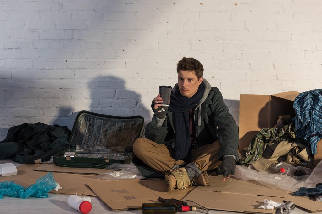 dakloze man houden papier beker zittend op karton omgeven door vuilnis - Foto, afbeelding