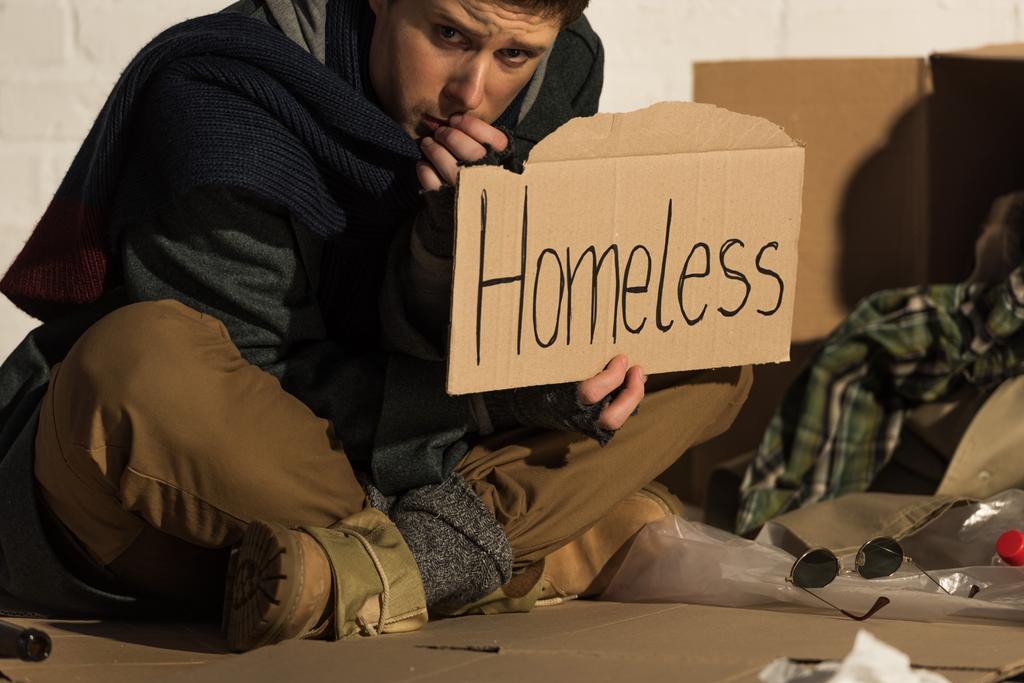 masentunut mies istuu pahvilla ja pitää kortin kanssa "koditon" käsin kirjoitettu teksti
 - Valokuva, kuva