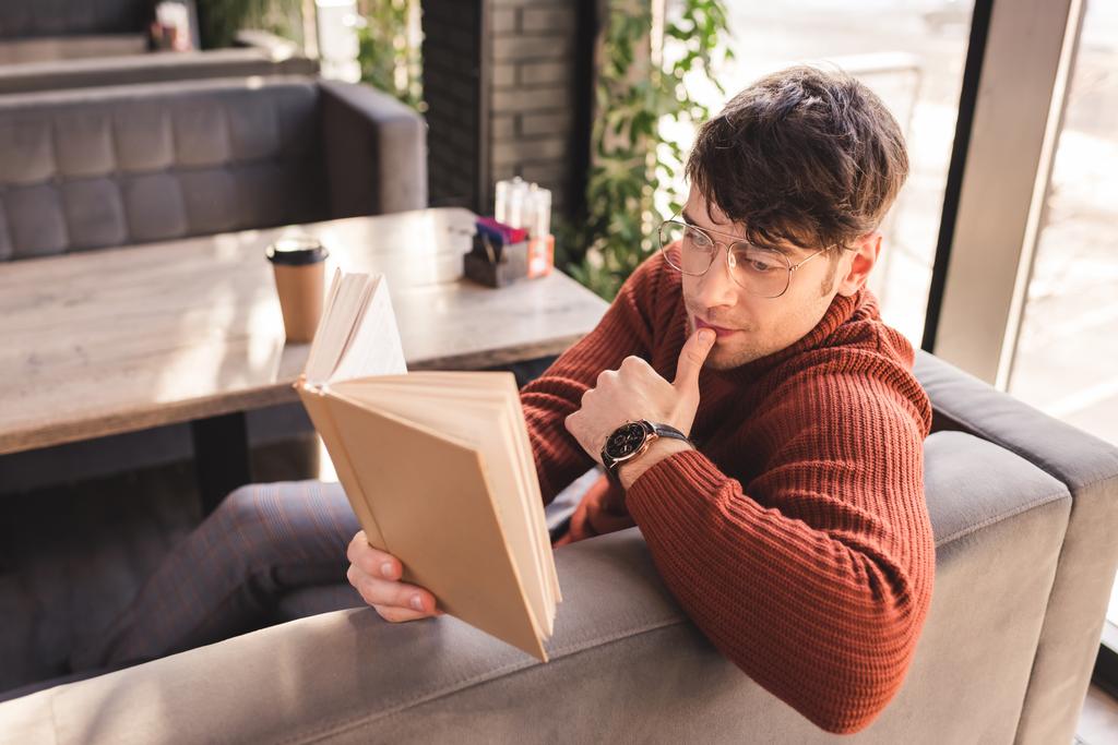 zamyšlený muž v brýlích, čtení knihy poblíž pohárek v kavárně - Fotografie, Obrázek