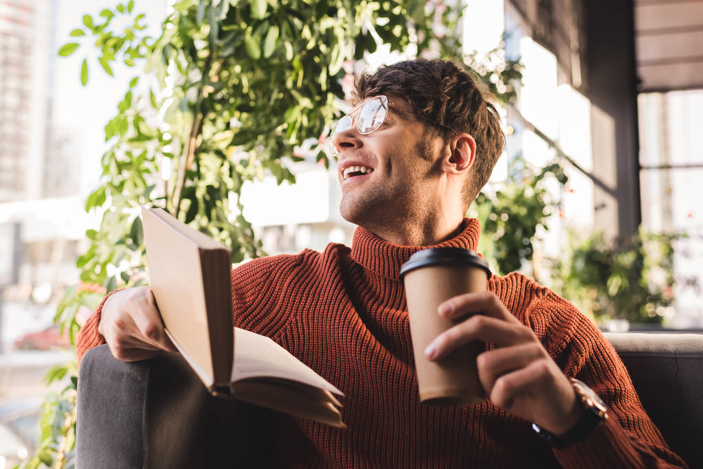 веселий молодий чоловік в окулярах тримає книгу і паперову чашку в руках
 - Фото, зображення