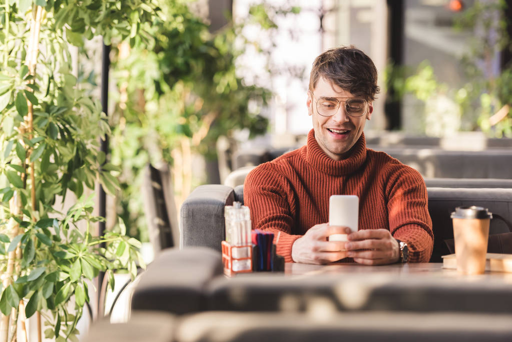 Selektiver Fokus eines lächelnden Mannes mit Smartphone in der Nähe von Einwegbecher und Buch im Café - Foto, Bild