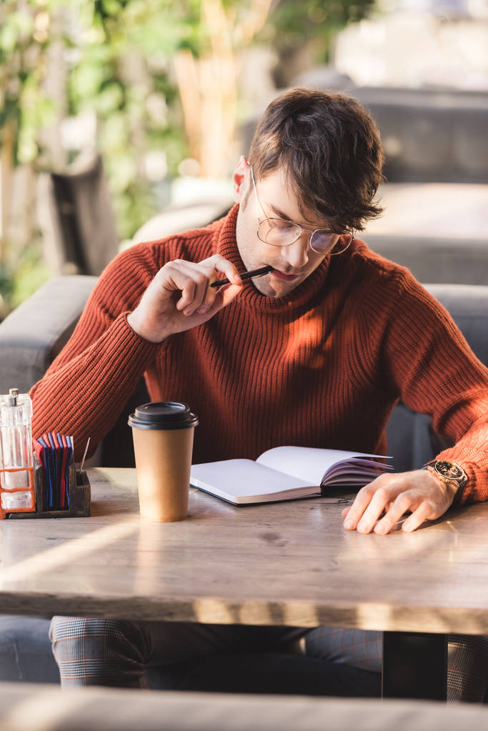 rozważny człowiek w okularach, trzymając pióro w pobliżu usta patrząc na notebook w pobliżu kubek jednorazowy w kawiarni - Zdjęcie, obraz