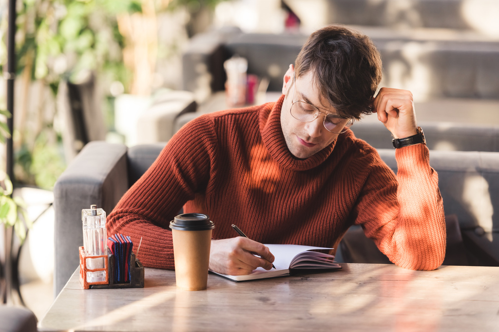 homme réfléchi dans des lunettes écrit dans un cahier près d'une tasse jetable dans un café
 - Photo, image