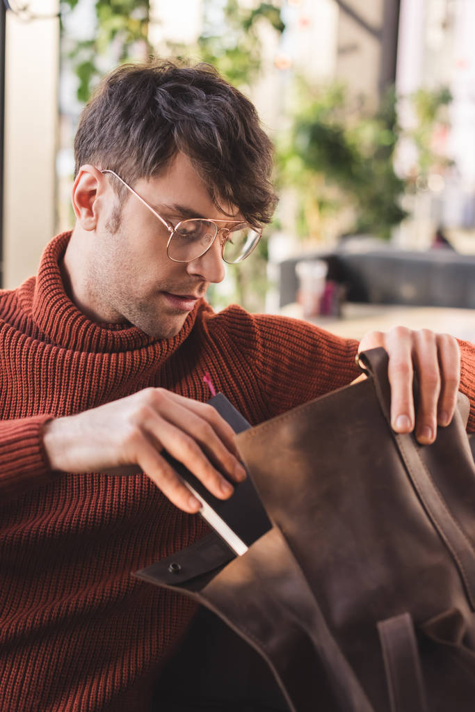 όμορφος άνδρας σε ποτήρια βάζοντας βιβλίο στην τσάντα σε καφέ - Φωτογραφία, εικόνα