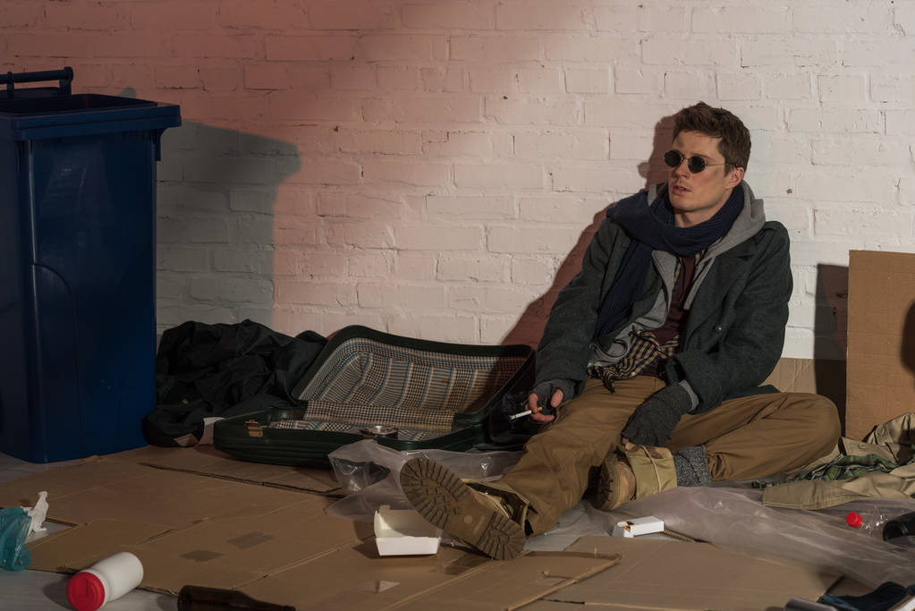 Obdachloser sitzt auf Müllhalde an Ziegelmauer und raucht Zigarette - Foto, Bild